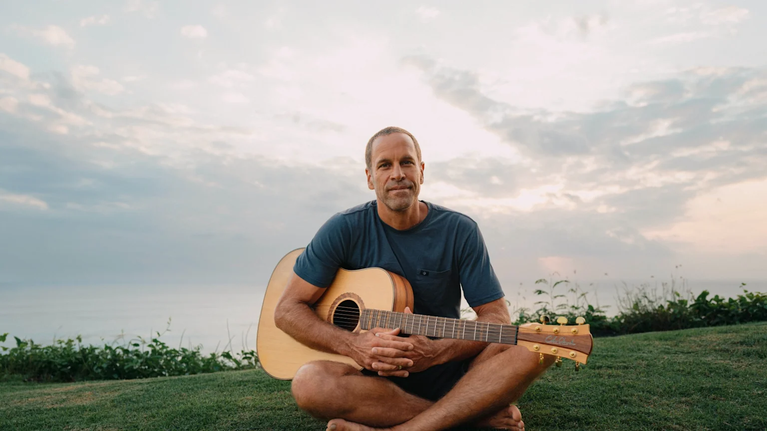 A man sits cross-legged on green grass, holding an acoustic guitar, with the ocean and a cloudy sky in the background.
