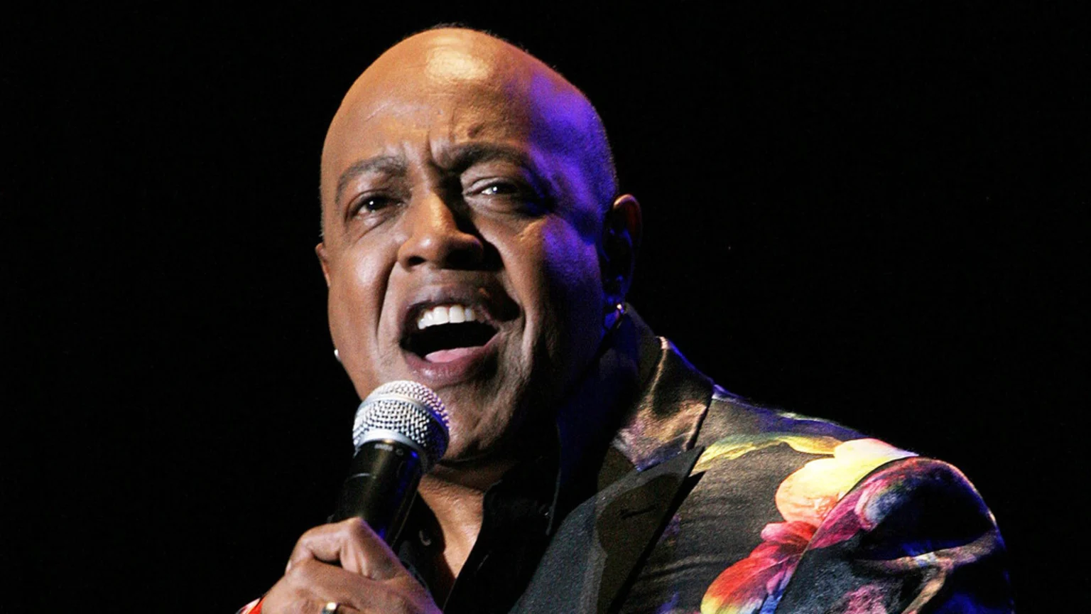 A bald, dark-skinned man with an open mouth sings into a microphone, wearing a black shirt and a dark blazer with a colorful floral pattern on a dark stage.