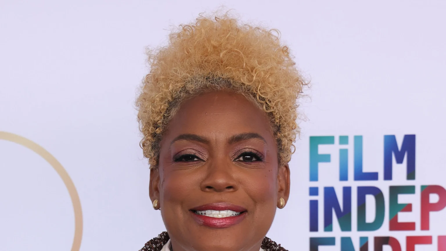 A smiling Black woman with short, curly blonde hair, dark pink makeup, and gold stud earrings stands against a white background with a partial golden circle and a blurred "FILM INDEP" logo.