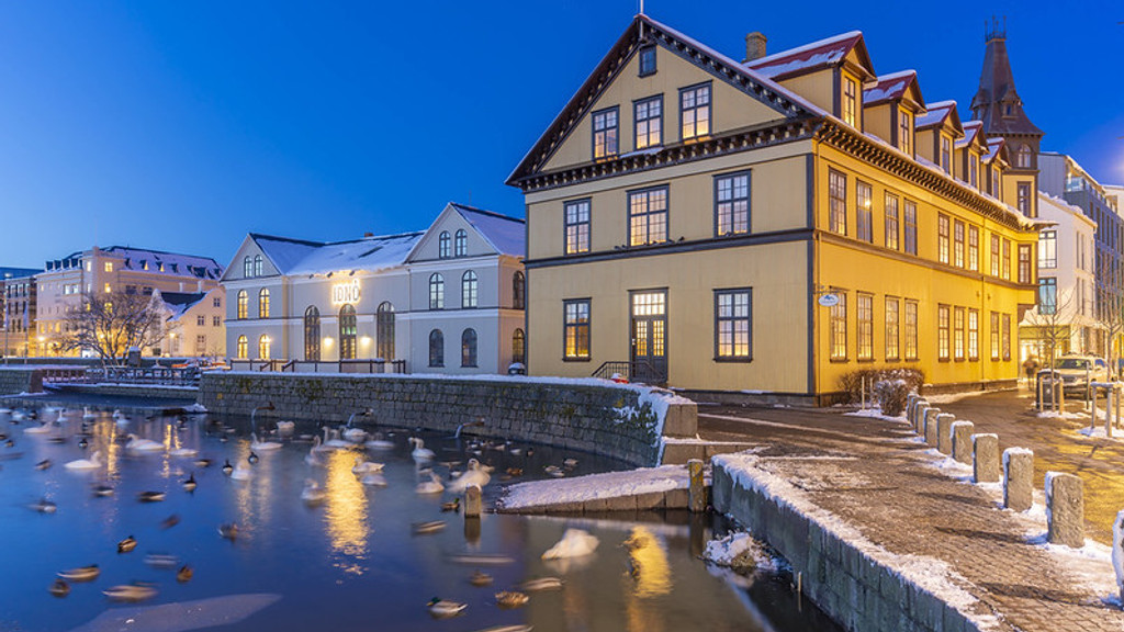 A blue hour winter cityscape with a prominent yellow building and other structures, a snow-covered canal with many swimming swans and ducks, and illuminated streets and windows.