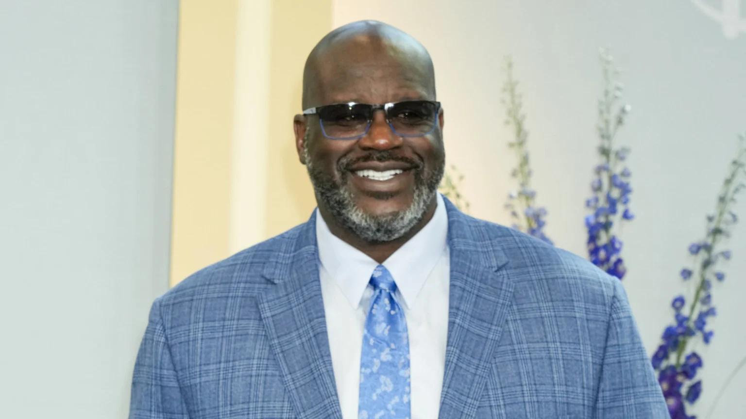 Shaquille O'Neal smiles at the camera, wearing blue-rimmed sunglasses, a blue plaid suit jacket, a white shirt, and a patterned blue tie, with a light, blurred background.