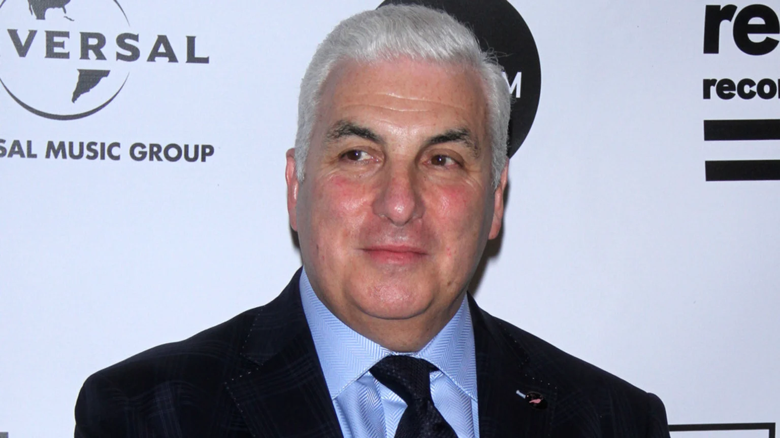 A man with grey hair wears a dark patterned suit, blue shirt, and dark tie, smiling slightly, with music group logos in the background.