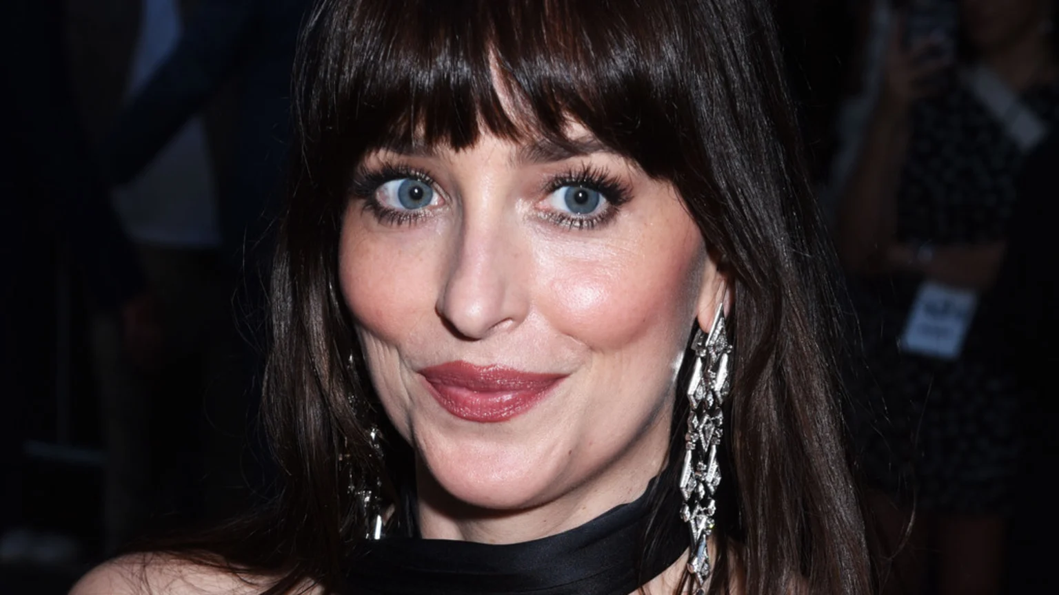 Close-up of actress Dakota Johnson smiling with wide blue eyes, dark hair and bangs, rosy cheeks, reddish-pink lips, and large dangling silver earrings against a dark background.