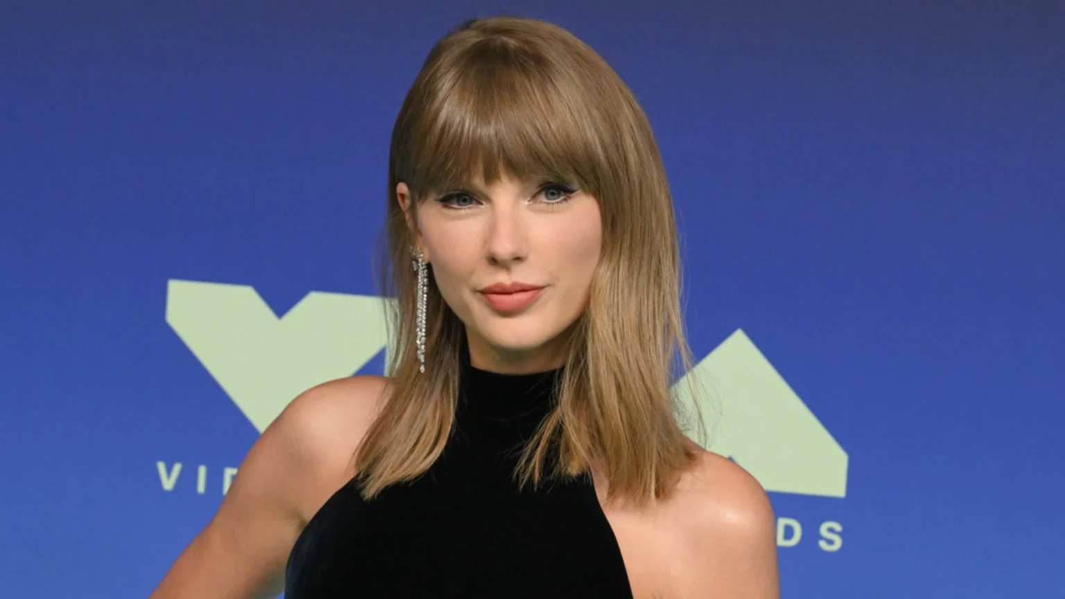 Taylor Swift, with light brown hair and bangs, smiles subtly at the camera, wearing a black top and long sparkly earrings, against a blue background with light green shapes.
