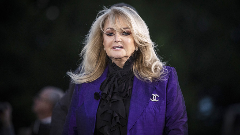 A close-up portrait of singer Bonnie Tyler, with long blonde hair, wearing a purple blazer, black ruffled blouse, and a sparkly Chanel brooch.