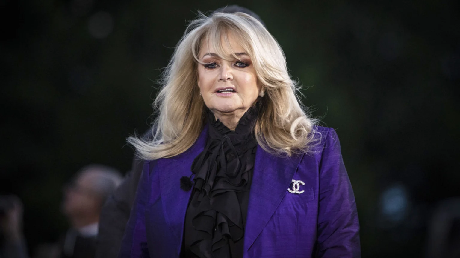 A close-up portrait of singer Bonnie Tyler, with long blonde hair, wearing a purple blazer, black ruffled blouse, and a sparkly Chanel brooch.