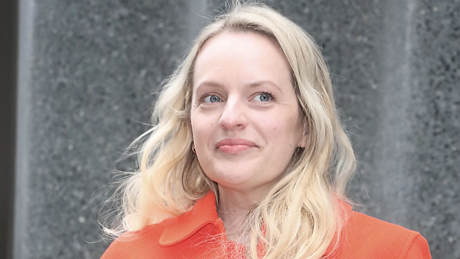 A smiling woman with blonde, wavy hair and blue eyes, wearing an orange collared top, looks slightly up and to the right against a blurry grey background.