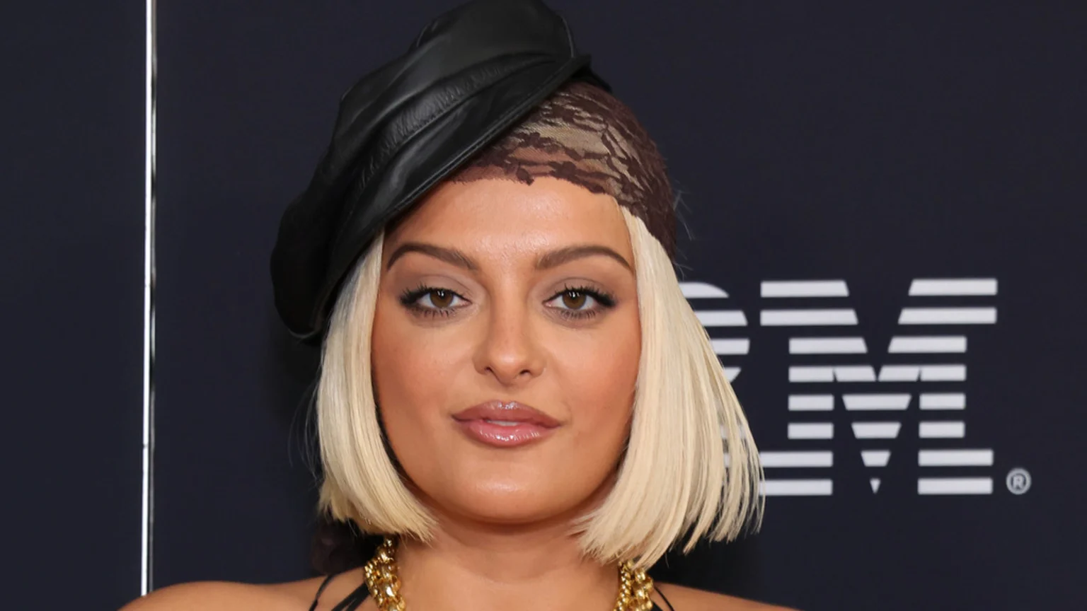 A close-up portrait of a woman with light blonde chin-length bob hair, brown eyes, and neutral expression. She wears a black leather-like beret-style head covering layered over a brown patterned head wrap, and a chunky gold-colored chain necklace. The background is dark with a white IBM logo and trademark symbol on the right, and a thin vertical white light strip on the left.