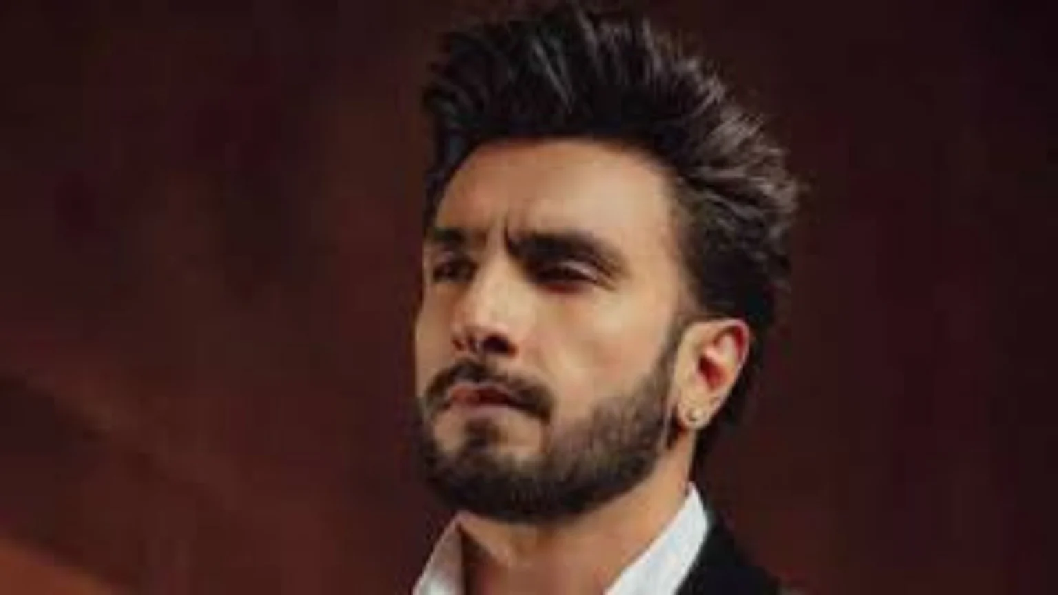 Ranveer Singh looks left with a serious expression, featuring styled dark hair, a beard, and an earring, wearing a white collared shirt and a dark jacket.