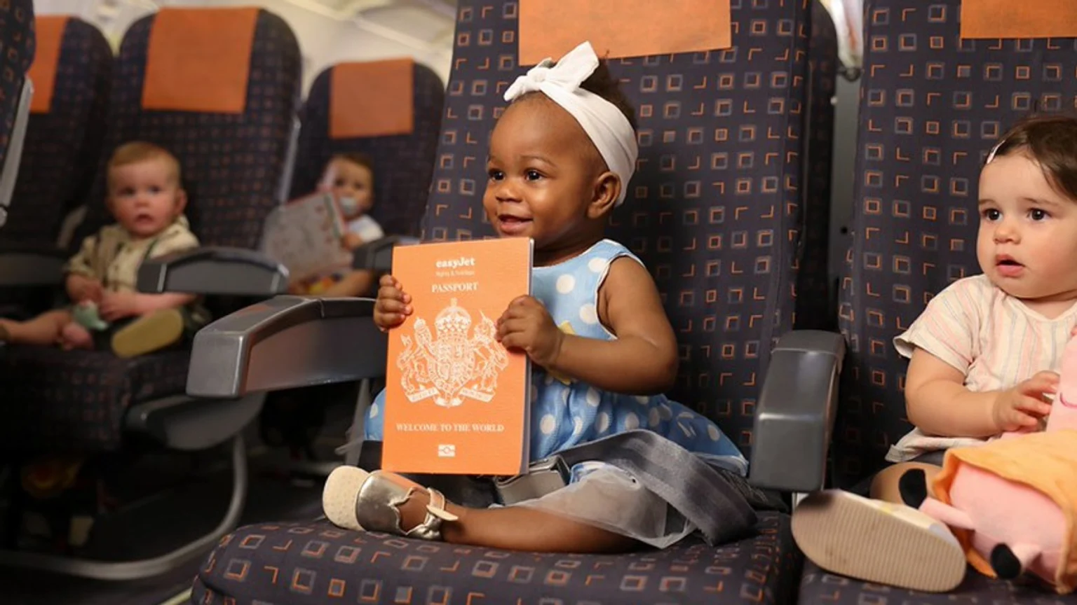 A baby smiles while holding an orange easyJet passport in an airplane seat, with other babies visible in the background.