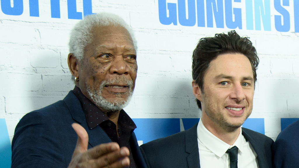 Morgan Freeman, with a grey beard and a gold earring, gestures while standing next to a smiling Zach Braff, who wears a suit and tie, against a white brick wall with blue text.