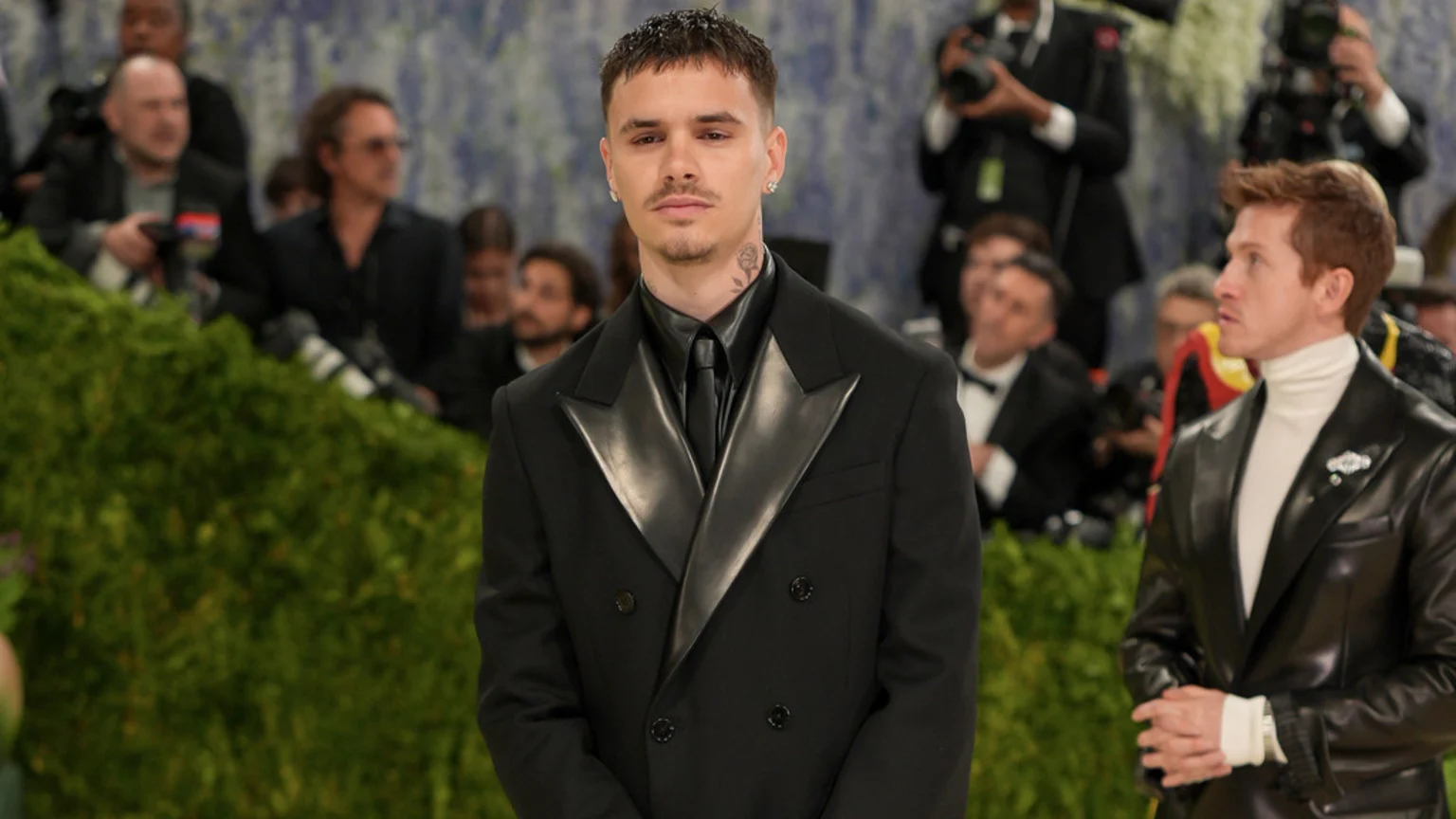 Romeo Beckham poses in a black double-breasted suit with shiny black lapels and a black tie at an event, with another man in a black leather jacket and white turtleneck next to him.