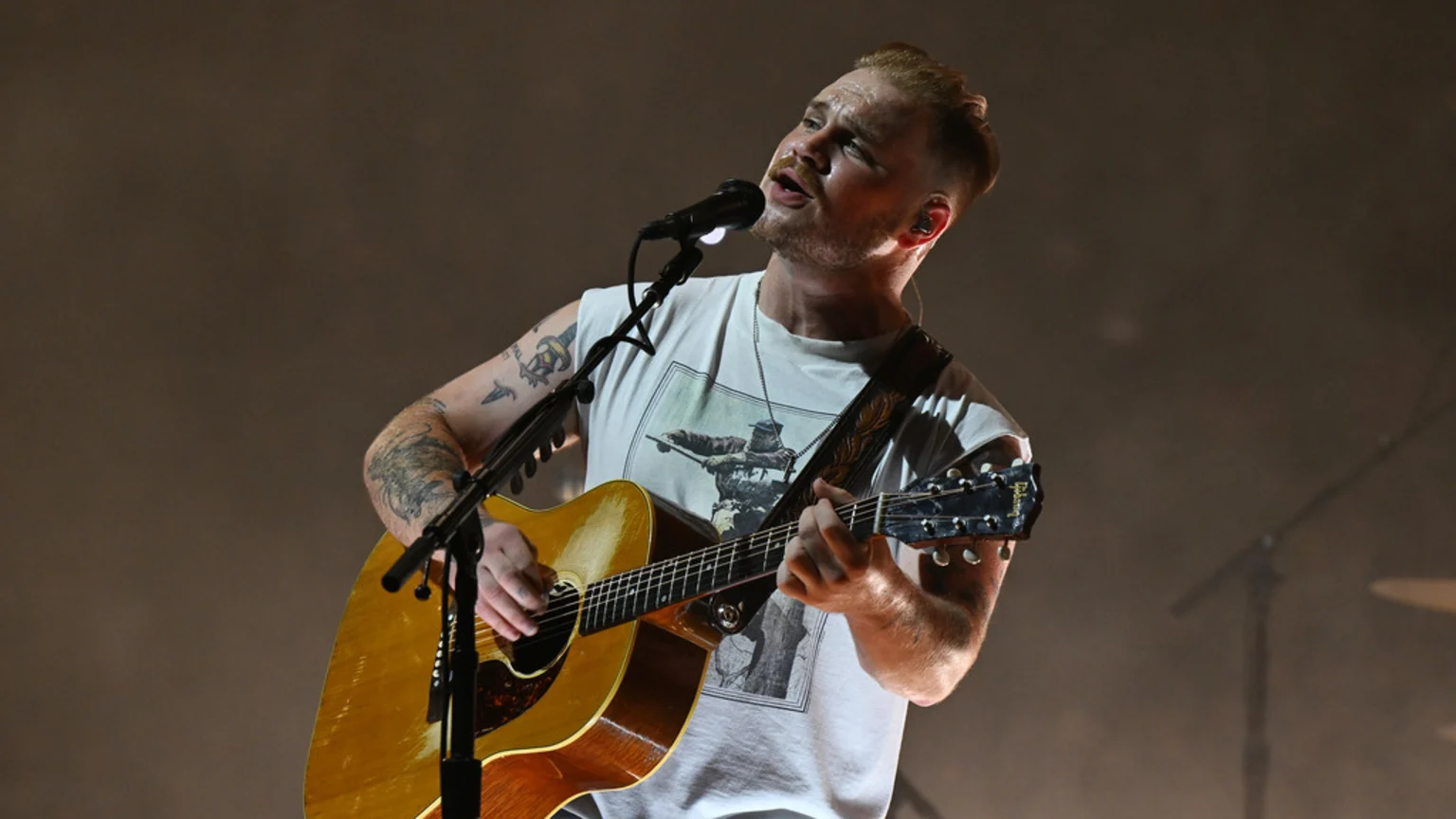 A male musician with a beard and tattoos sings into a microphone while playing an acoustic guitar on stage.