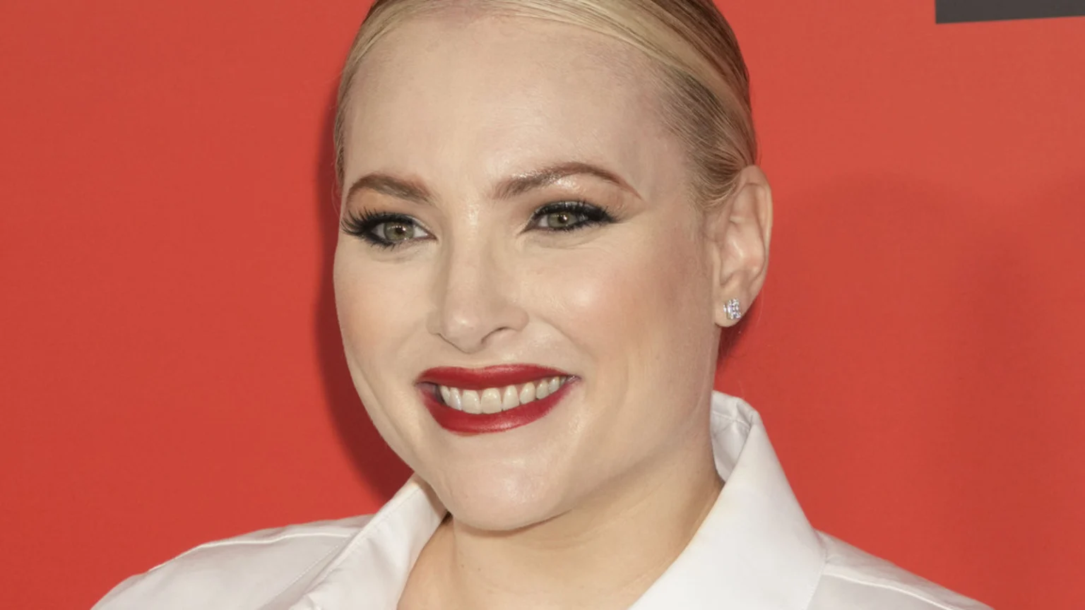 Close-up of Meghan McCain smiling, with blond hair pulled back, heavy eye makeup, red lipstick, and diamond stud earrings, against a red background.