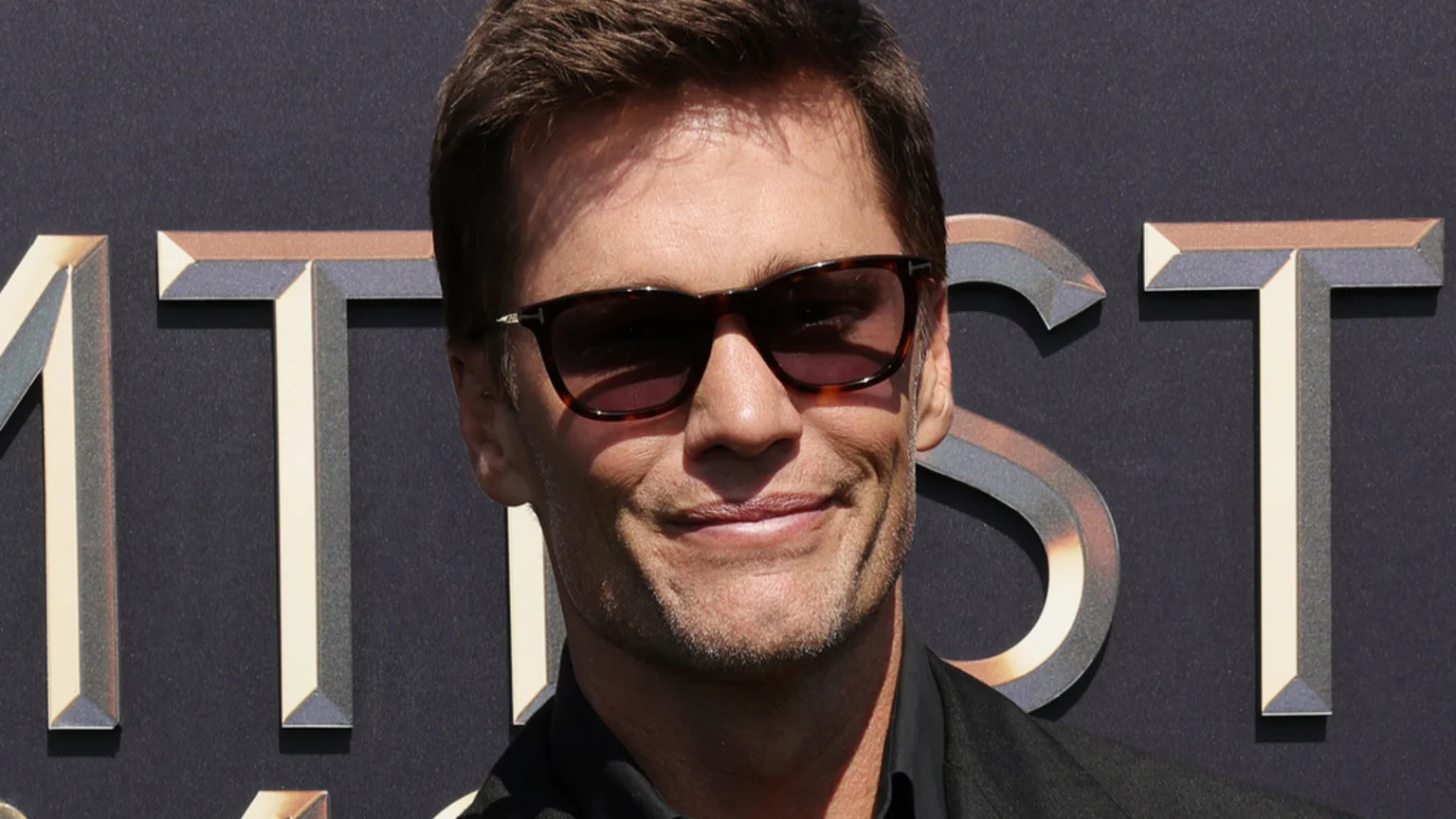 Close-up of Tom Brady smiling and wearing dark sunglasses against a dark background with stylized metallic letters.
