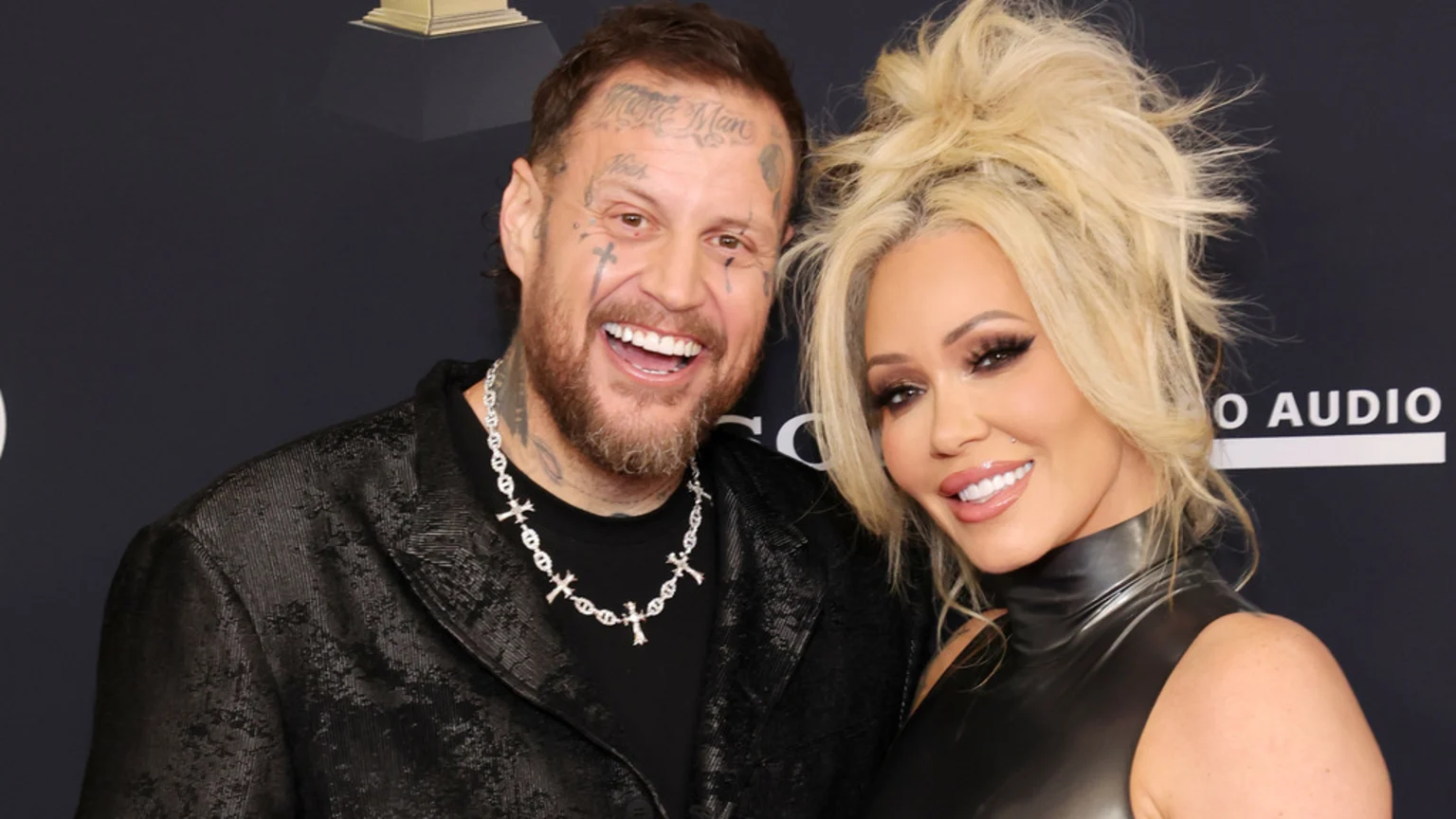 A smiling man with facial tattoos and a beard, wearing a cross necklace and black jacket, poses next to a smiling woman with blonde hair, heavy makeup, and a black high-necked top.