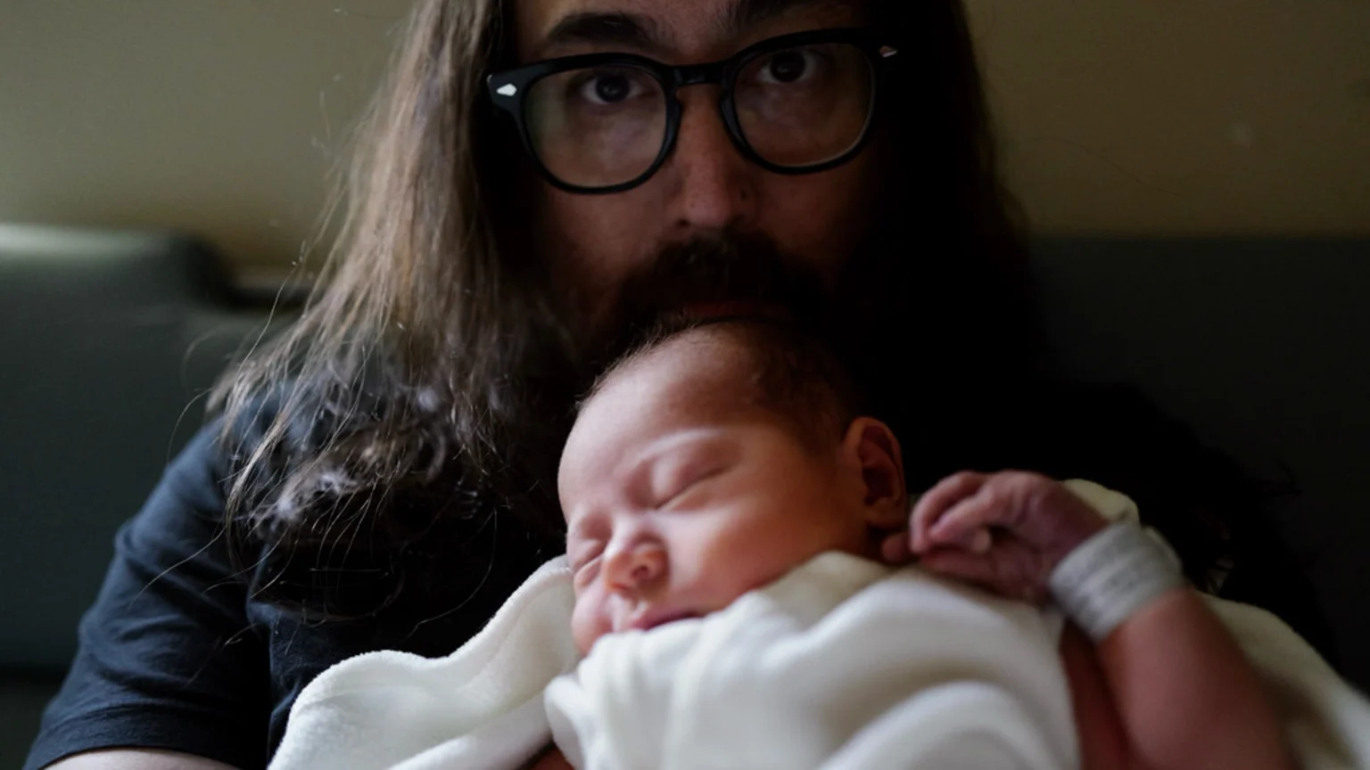 A man with long dark hair, a beard, and glasses looks at the camera while holding a sleeping newborn baby wrapped in a white blanket, wearing a hospital wristband.