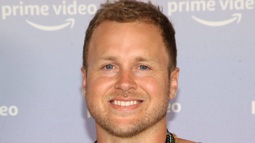 A close-up headshot of a smiling man with light brown hair, blue eyes, and a short beard. He is grinning broadly, showing his teeth, against a light blue background featuring blurred "prime video" logos.