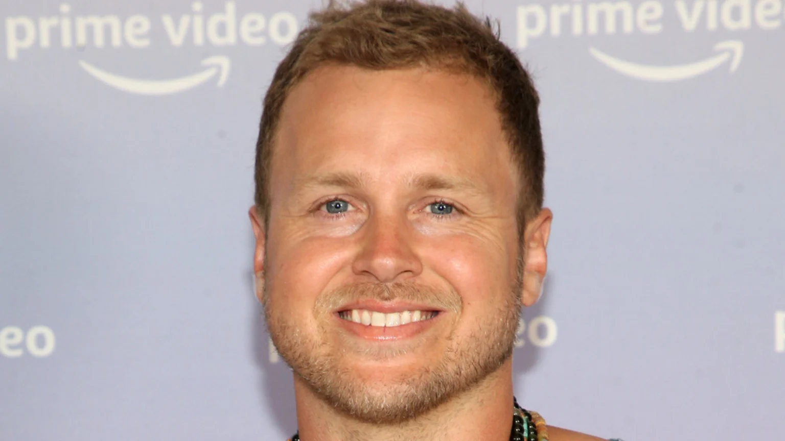 A close-up headshot of a smiling man with light brown hair, blue eyes, and a short beard. He is grinning broadly, showing his teeth, against a light blue background featuring blurred "prime video" logos.