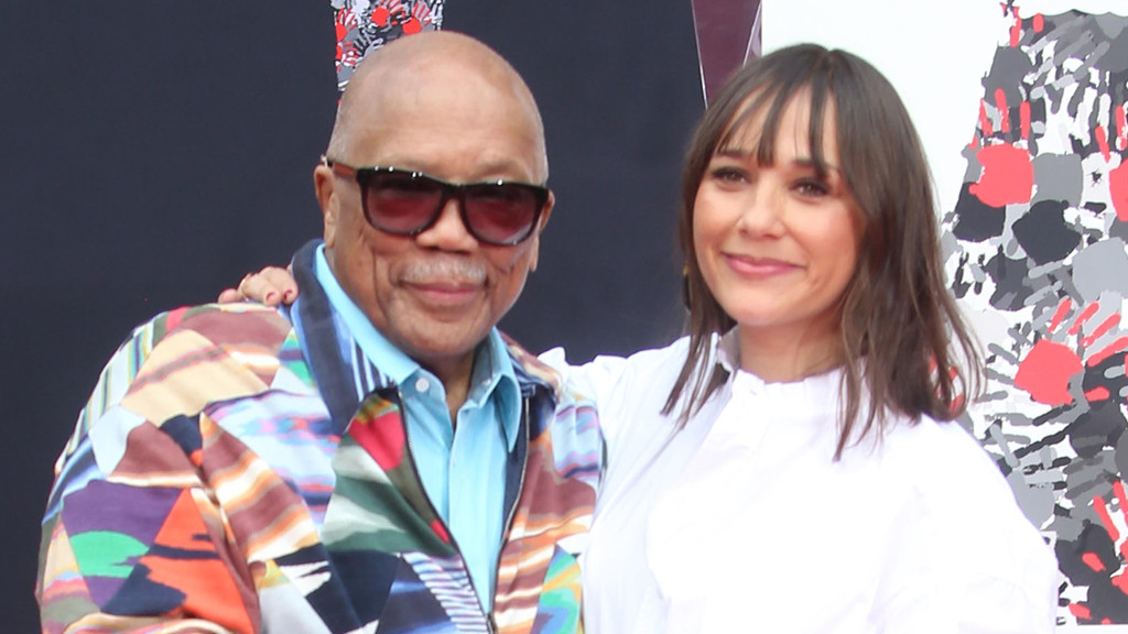 Quincy Jones, bald with sunglasses and a colorful jacket, smiles next to a smiling woman with dark hair and bangs in a white shirt, with her hand on his shoulder.