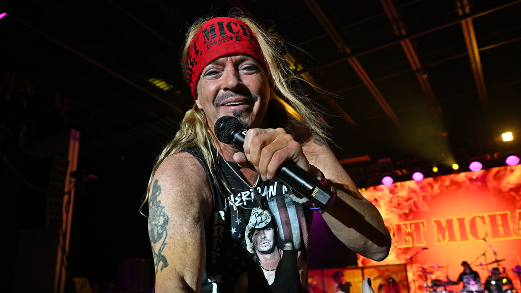 Bret Michaels, with long blonde hair and a red bandana, smiles while holding a microphone on stage, wearing a black tank top, with a "BRET MICHAELS" fiery backdrop.