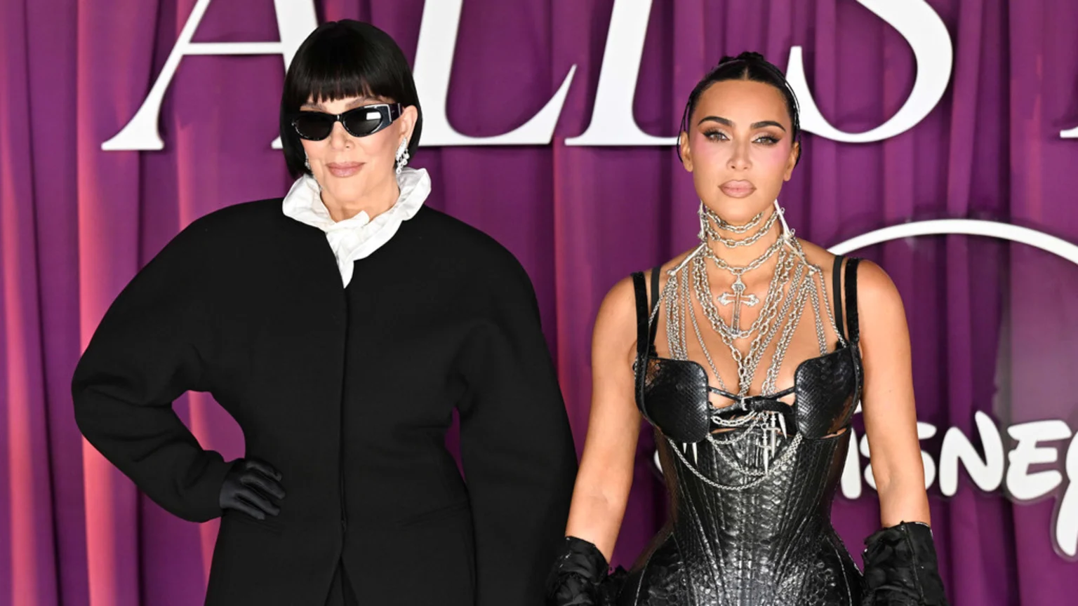 Kris Jenner in a black coat and white ruffled shirt stands next to Kim Kardashian in a black corset and layered silver chains, both posing against a purple backdrop.