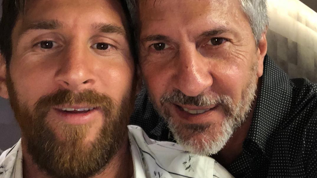 A close-up selfie of Lionel Messi, with a brown beard, smiling next to an older man with grey hair and a grey beard, both looking forward.