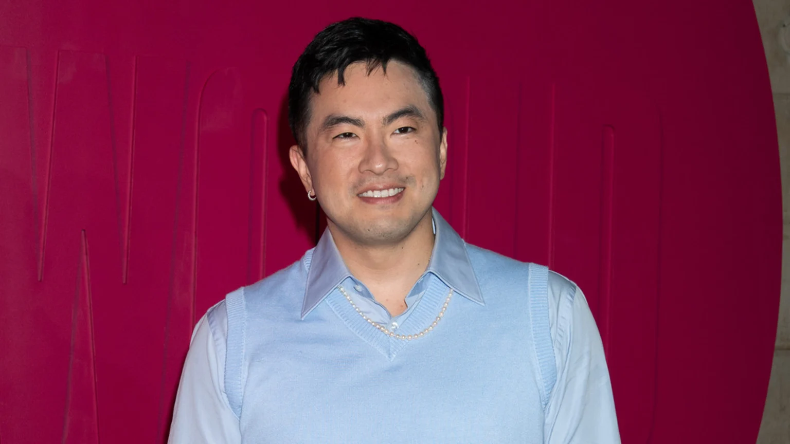 Bowen Yang smiles, wearing a light blue collared shirt, a matching sweater vest, and a pearl necklace, against a textured magenta background.