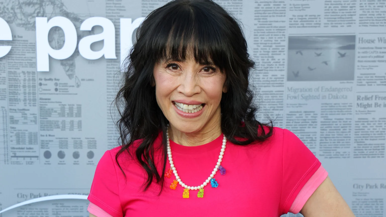 Actress Lauren Tom smiles broadly, wearing a bright pink shirt and a pearl necklace with gummy bear charms, against a blurred background with newspaper-like text.