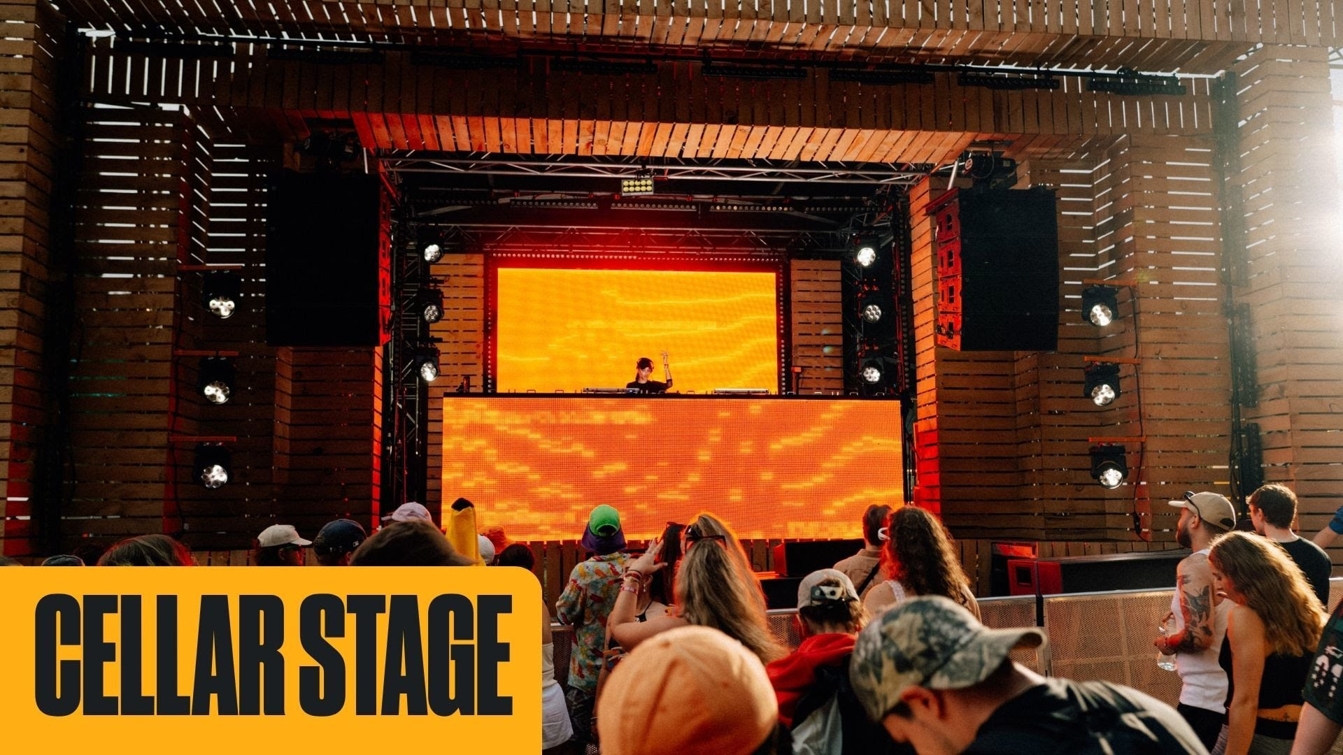 A DJ performs on a wooden outdoor stage featuring large orange LED screens, with a crowd watching, and a "CELLAR STAGE" banner in the foreground.