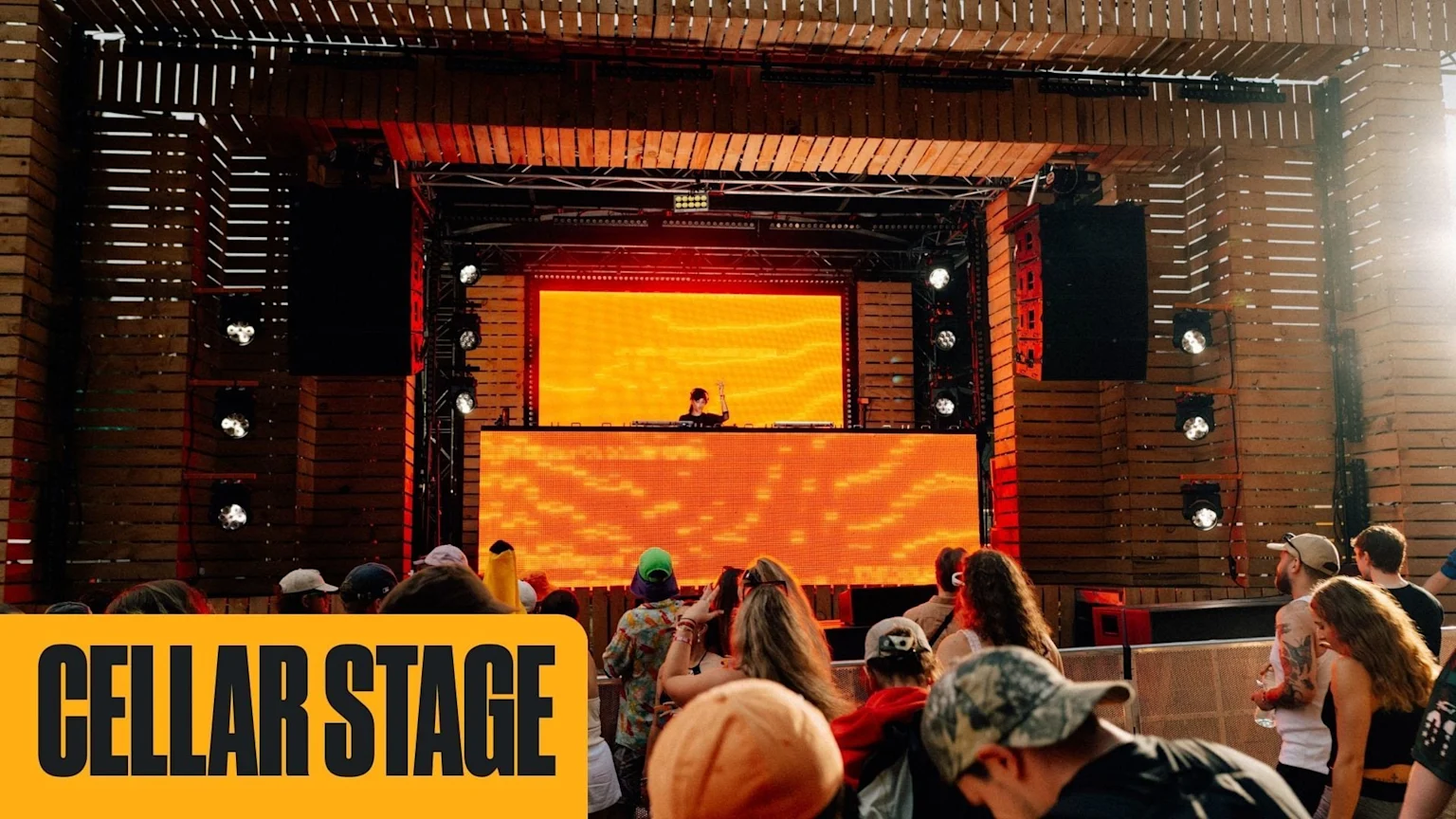 A DJ performs on a wooden outdoor stage featuring large orange LED screens, with a crowd watching, and a "CELLAR STAGE" banner in the foreground.