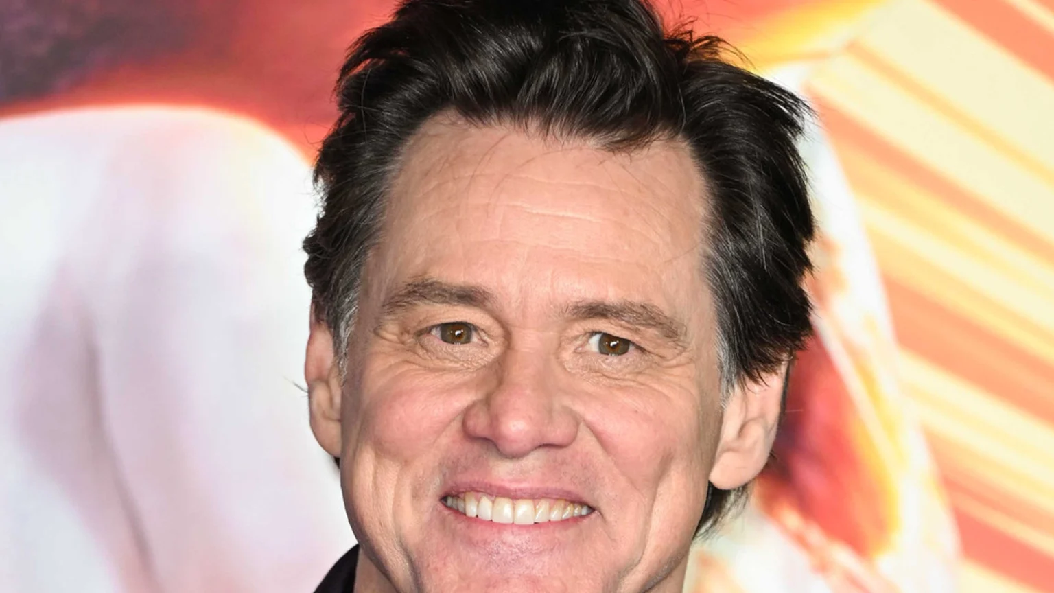 A close-up shot of actor Jim Carrey smiling, showing his teeth, with dark hair, some graying at the temples, and visible wrinkles on his face, against a blurred, warm-colored background.