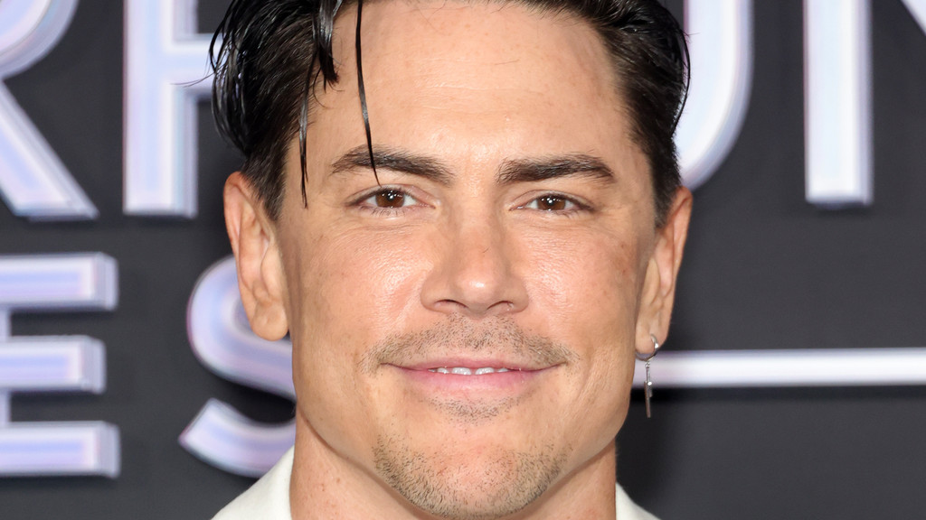 A close-up of Tom Sandoval smiling directly at the camera, featuring his slicked-back dark hair, brown eyes, mustache, and chin stubble, with a silver earring visible.
