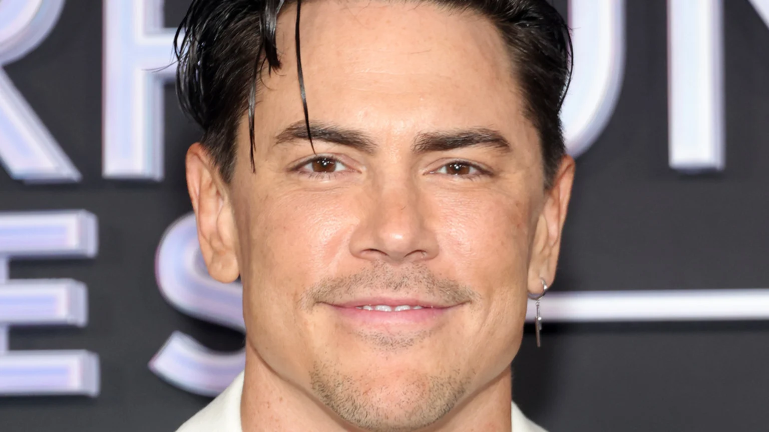 A close-up of Tom Sandoval smiling directly at the camera, featuring his slicked-back dark hair, brown eyes, mustache, and chin stubble, with a silver earring visible.