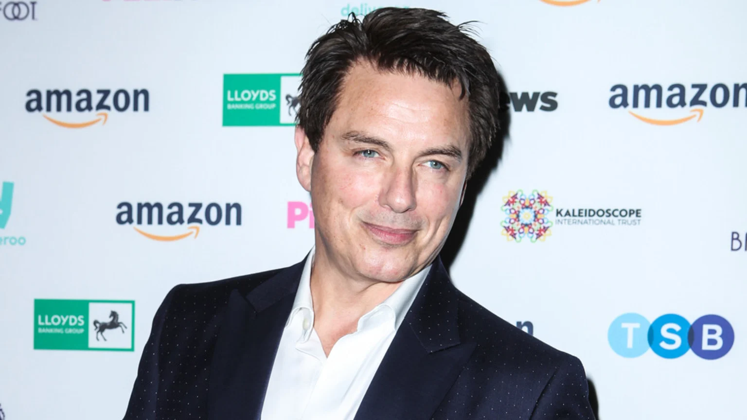 John Barrowman, with dark hair and blue eyes, smiles slightly in a dark suit with white dots, standing against a white backdrop featuring various company logos.