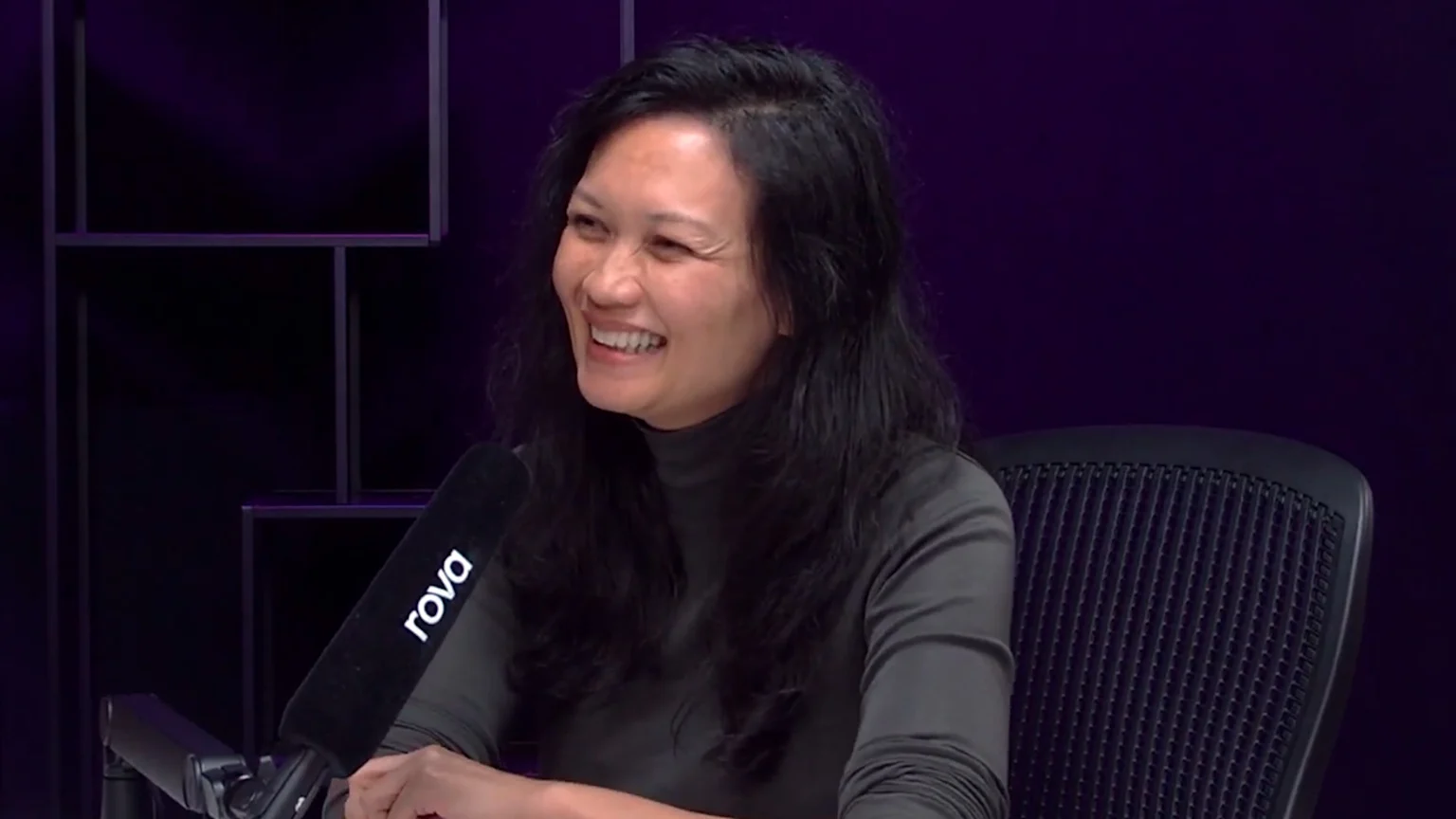 A joyful East Asian woman with long dark hair and a dark grey turtleneck smiles broadly while seated in an indoor podcast studio. A black microphone labeled 'rova' is in the foreground, with a black office chair and dark purple studio background behind her.