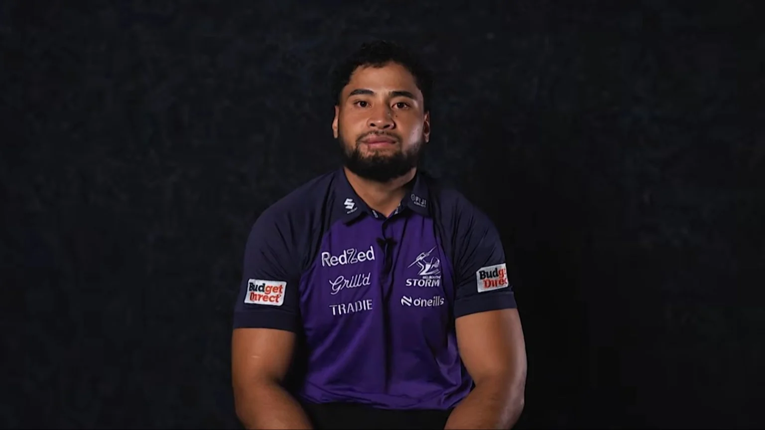 A man with a beard and dark hair, wearing a purple and dark blue Melbourne Storm polo shirt, looks directly at the viewer against a dark background.
