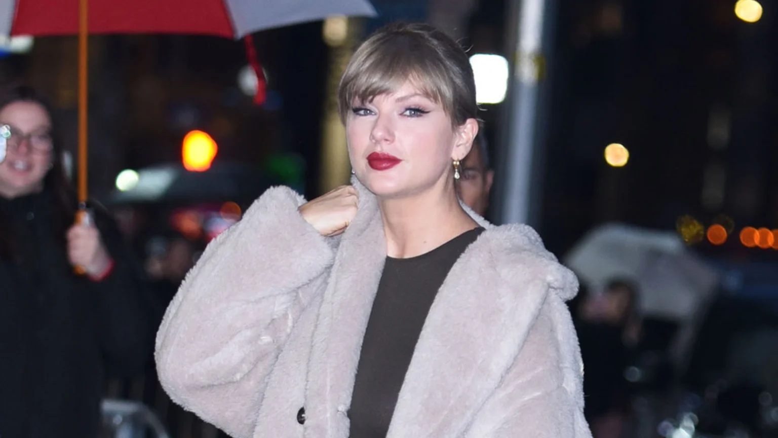 A candid night-time portrait of Taylor Swift wearing a light beige voluminous faux fur coat and red lipstick. She is adjusting her coat, with winged eyeliner and gold drop earrings visible. Blurred figures and city lights form the background.