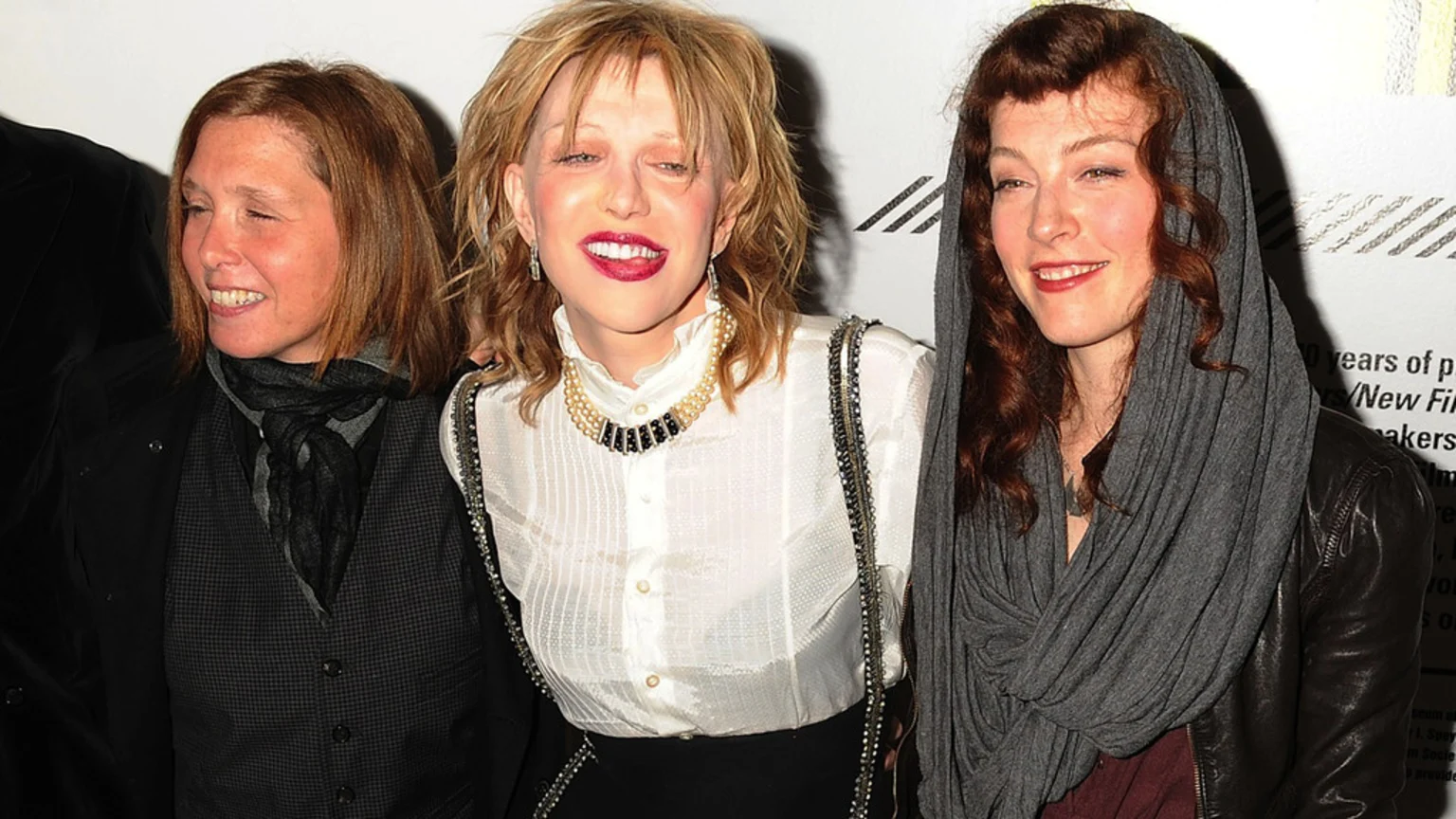 Courtney Love smiles in a white blouse and pearls between Melissa Auf der Maur in a dark vest and scarf, and a woman with red hair wearing a grey headscarf and leather jacket.