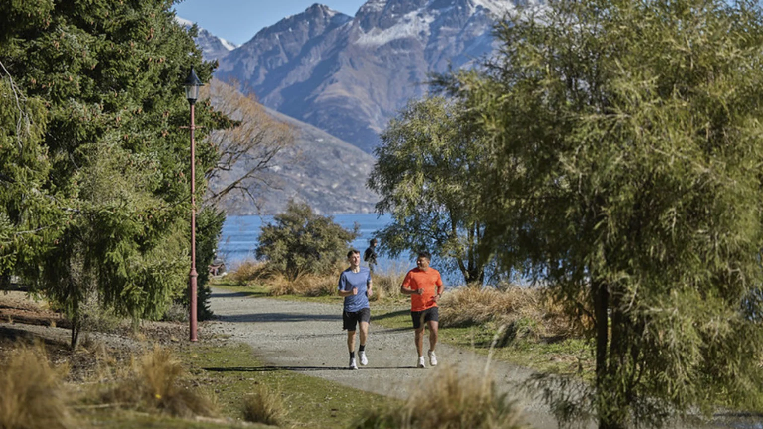 Two men jog on a scenic path alongside a lake with snow-capped mountains in the background, suggesting an active outdoor adventure.