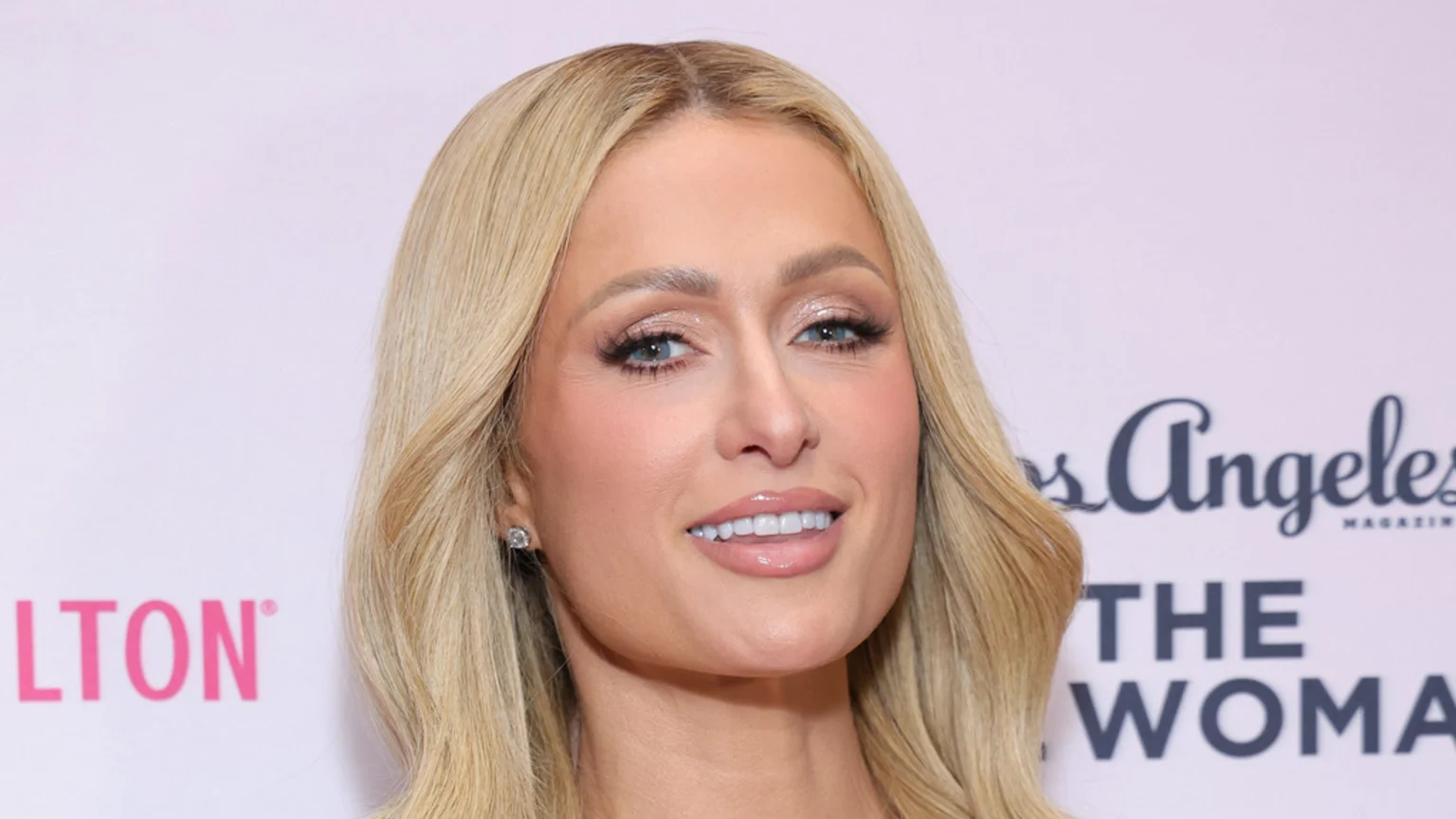 A close-up of Paris Hilton smiling slightly, showing her teeth, with long blonde hair, bright blue eyes, soft makeup, and a diamond stud earring, against a light background with blurry text.