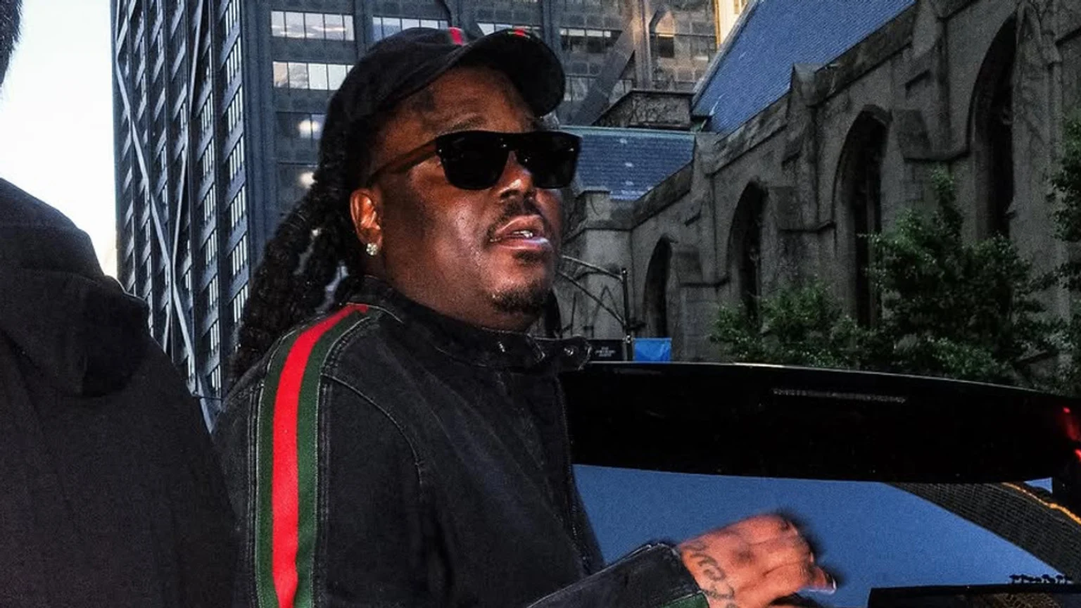A dark-skinned man with dreadlocks, wearing sunglasses, a black cap, and a black jacket with a red and green vertical stripe, stands outdoors with city buildings behind him.