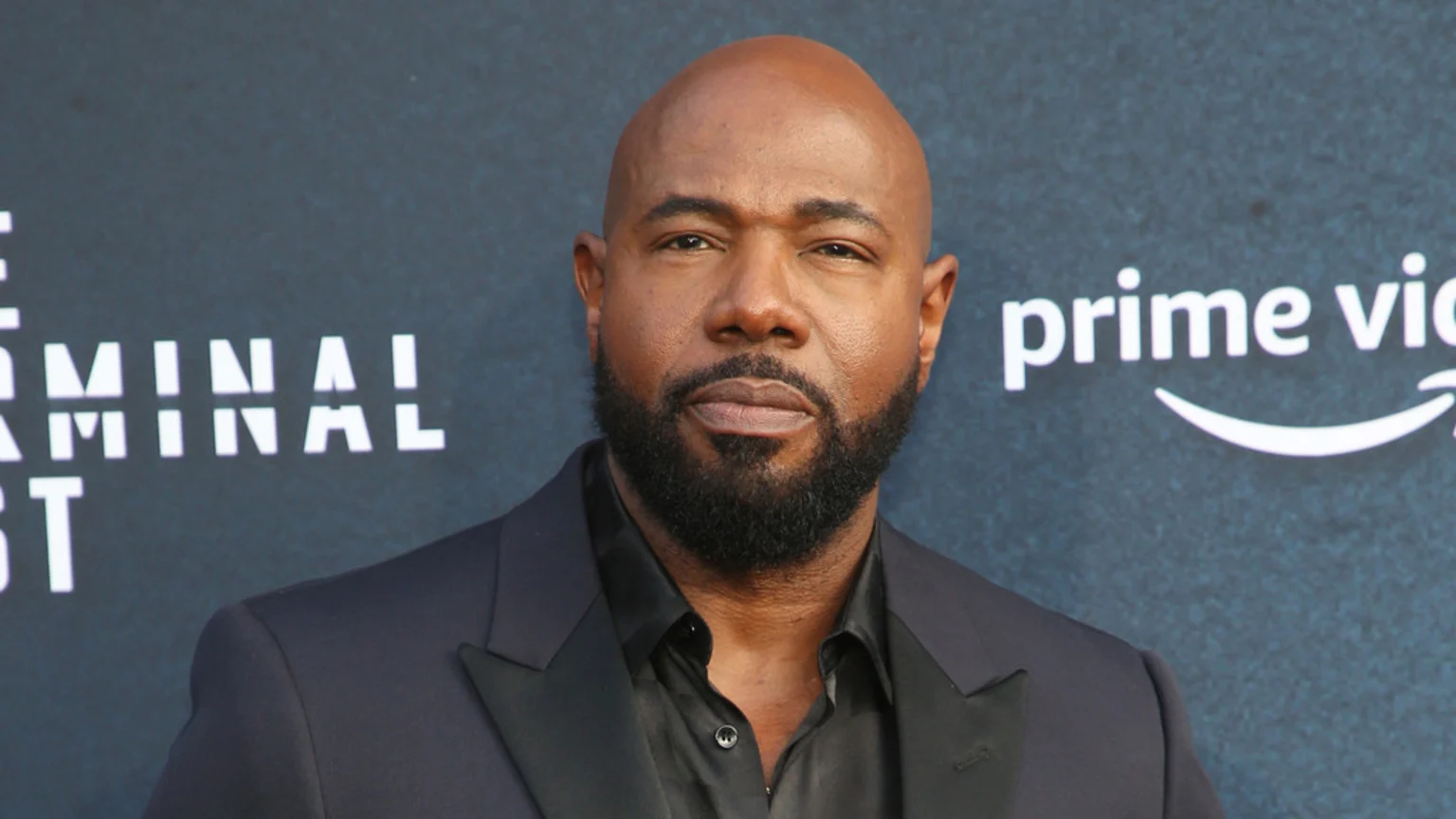 A bald man with a dark beard and dark skin wears a dark suit and shirt, standing in front of a dark background with "prime video" and "TERMINAL LIST" visible.