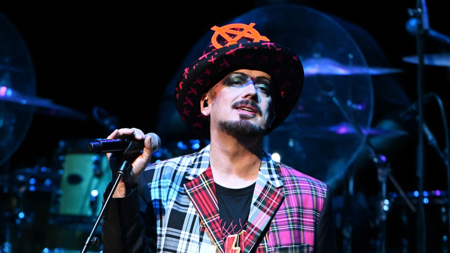 Boy George, wearing a distinctive hat with an orange anarchy symbol, stage makeup, and a colorful plaid jacket, sings into a microphone on a dark stage with blurry blue lights.