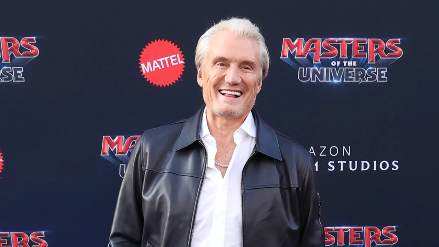 Dolph Lundgren smiles broadly, wearing a white shirt, silver chain, and black leather jacket, with "Mattel" and "Masters of the Universe" logos on the dark background.