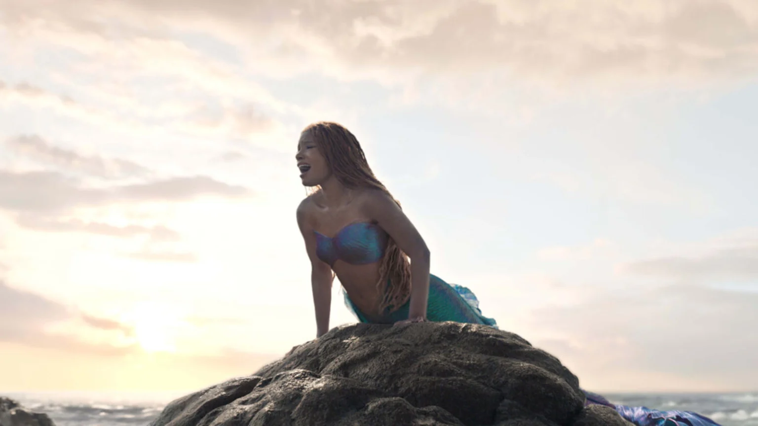 Halle Bailey as Ariel, with reddish-brown dreadlocks and a shimmering blue-purple top and green tail, on a dark rock against a bright, cloudy ocean sunset, appearing to sing.