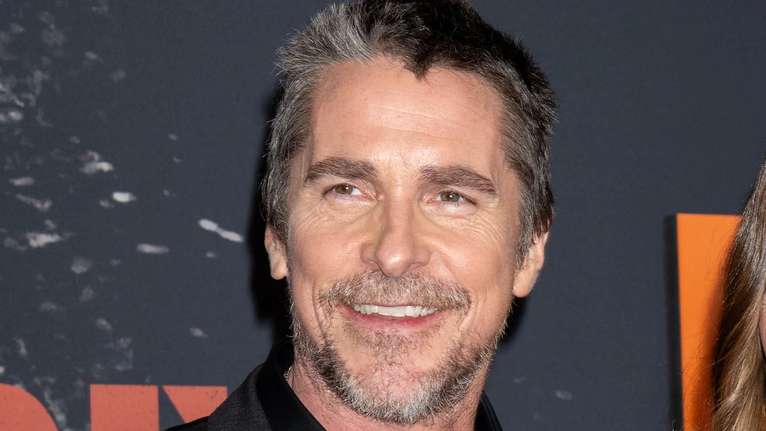 Christian Bale smiles, revealing his teeth, with graying hair and a beard, against a dark background with subtle texture.
