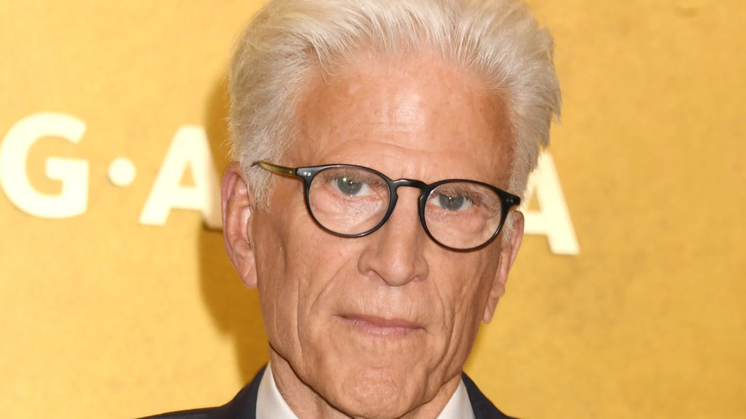 Close-up portrait of an older man with white hair and dark-rimmed glasses, wearing a suit, against a blurred yellow background with white letters.