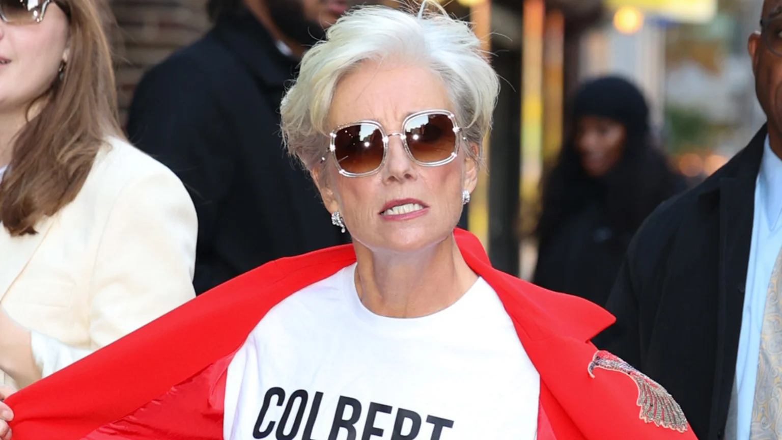 A woman with short grey hair and large sunglasses wears a white "COLBERT" t-shirt and a red blazer, looking a bit perturbed, with blurred people in the background.