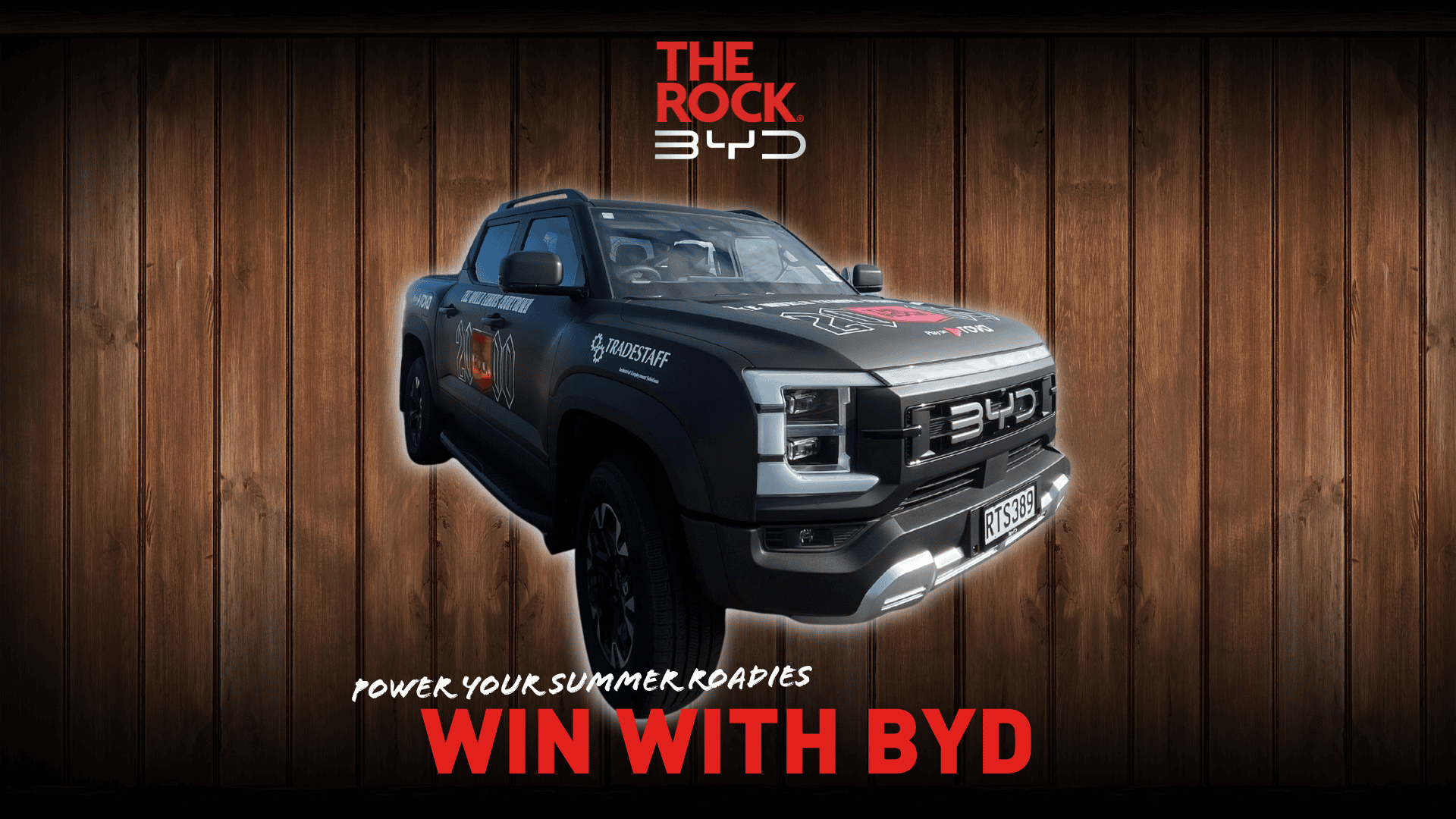 A black BYD branded pickup truck with "The Rock" and "Tradestaff" logos is angled on a wooden background, with text "Power Your Summer Roadies Win With BYD" below.
